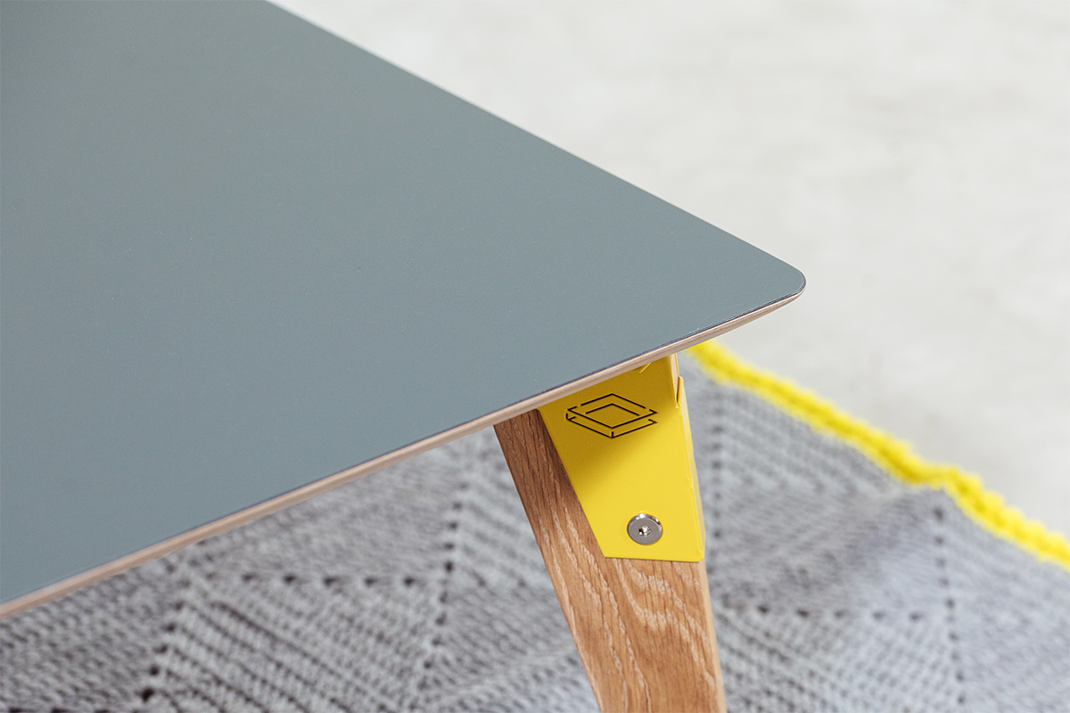 corner detail of modpop coffee table with grey Fenix table top, yellow steel brackets and solid oak legs with grey and yellow rug in background